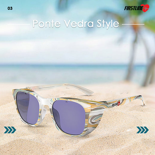 Safety goggles with gold and white frame on sand with ocean and palm tree in the background, branded 'Firstline'.